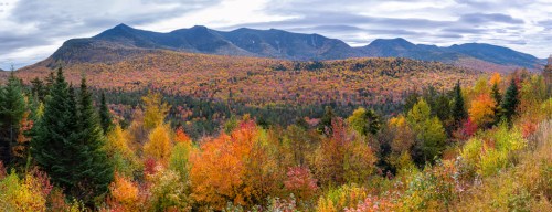 A panoramic view of mountains surrounded by vibrant autumn foliage in shades of orange, red, and yellow under a cloudy sky.