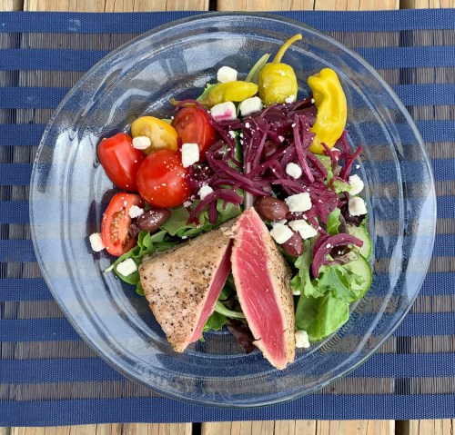 A glass plate with seared tuna, mixed greens, cherry tomatoes, olives, pickled onions, and feta cheese.