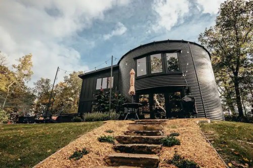 A modern, round black house surrounded by greenery, with a stone pathway leading to it under a cloudy sky.