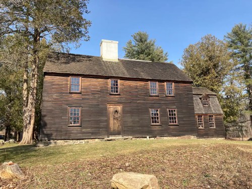 A historic wooden house with a steep roof, surrounded by trees and a clear blue sky.