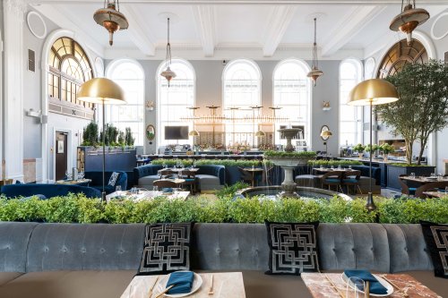 Elegant restaurant interior featuring plush seating, greenery, large windows, and stylish lighting fixtures.