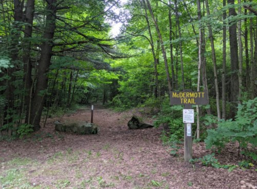 A wooded trail entrance marked "McDermott Trail," surrounded by lush green trees and rocky terrain.