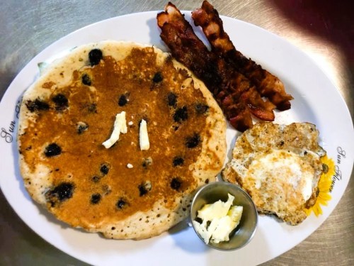 A plate with a large blueberry pancake, crispy bacon strips, and a fried egg with butter on the side.
