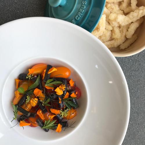 A white bowl with colorful tomatoes and edible flowers, alongside a bowl of light, fluffy snacks.