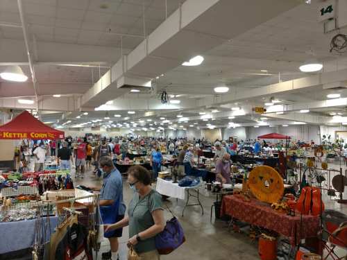 A bustling indoor market with vendors and shoppers, featuring various booths and tables filled with goods.