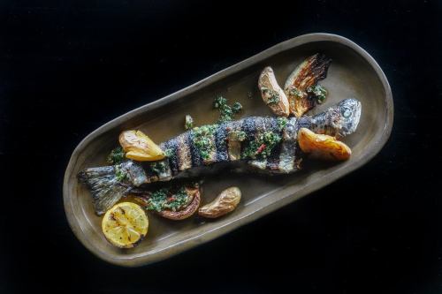 Whole grilled trout on an oblong plate with fennel, potato, salsa verde, and grilled lemon