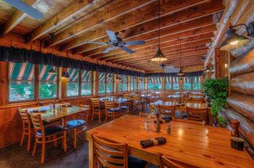Cozy wooden restaurant interior with tables, chairs, and large windows, featuring rustic decor and natural light.