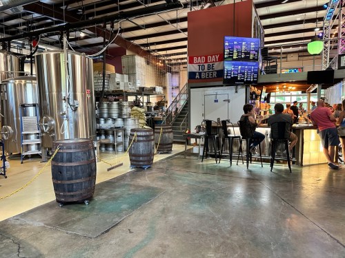 A brewery interior featuring large brewing tanks, a bar area with patrons, and wooden barrels in a spacious setting.