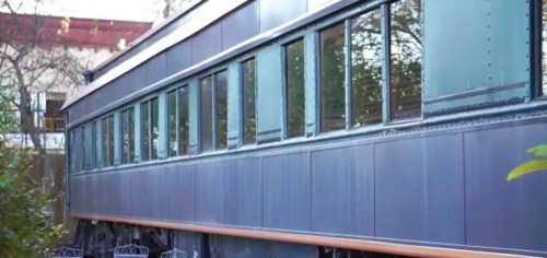 Side view of a vintage train car with multiple windows, surrounded by greenery.