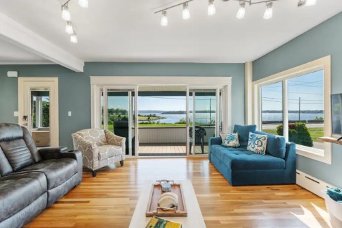 Bright living room with large windows overlooking a scenic view, featuring a blue sofa, armchair, and wooden flooring.