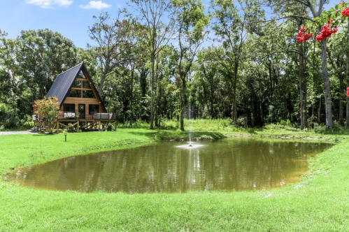 A scenic view of an A-frame cabin beside a pond, surrounded by lush greenery and trees under a clear blue sky.