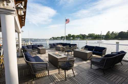 Outdoor seating area by the water, featuring sofas and tables, with a view of boats and a flag in the background.