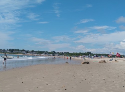 A sunny beach scene with people swimming, surfing, and relaxing on the sand under a blue sky with scattered clouds.