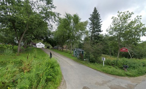 A gravel road lined with trees leads to a house and a small green shed, with a sign visible in the background.