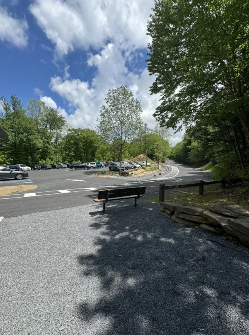A gravel path leads to a parking area surrounded by trees and a blue sky with clouds. A bench is visible in the foreground.