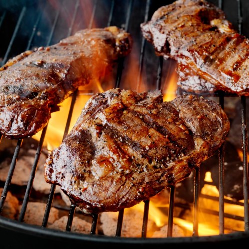 Grilled steaks sizzling on a barbecue grill with flames and smoke in the background.