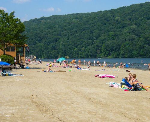 A sandy beach with people sunbathing and swimming, surrounded by green hills and a calm lake.