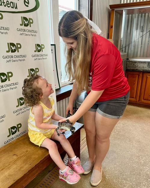 A young girl in a yellow dress smiles as a woman in a red shirt shows her a small animal at Gator Chateau.