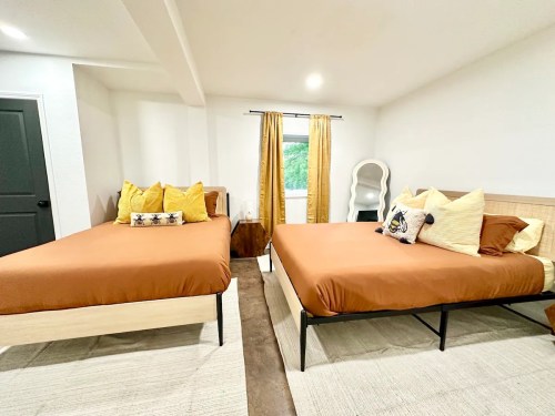 A bright, modern bedroom featuring two beds with brown bedding, yellow pillows, and large windows with curtains.
