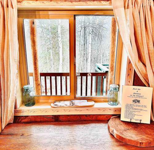 A cozy window view with wooden accents, jars, and a menu on a rustic table, overlooking a snowy forest.