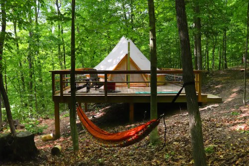 A glamping tent on a wooden deck surrounded by trees, with a hammock nearby in a serene forest setting.