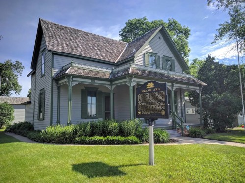 Historic house with green trim and a plaque for Sinclair Lewis, surrounded by grass and trees under a blue sky.