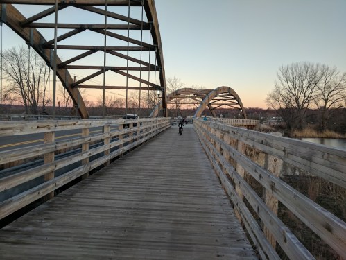 A wooden bridge with arched supports, leading into the distance at sunset, with a cyclist riding along the path.