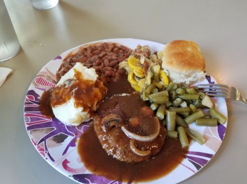 A plate of food featuring meatloaf with gravy, mashed potatoes, green beans, baked beans, corn casserole, and a biscuit.