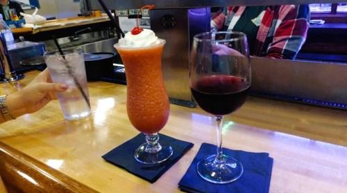 A colorful frozen drink with a cherry and a glass of red wine on a bar counter, with a hand holding a water glass.