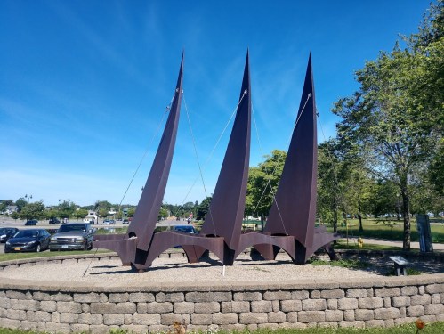 A large, abstract metal sculpture resembling three sails, set on a stone base in a park with trees and blue sky.