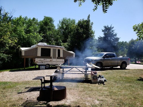 A pop-up camper and a truck are parked at a campsite with a picnic table and a fire pit, surrounded by trees.