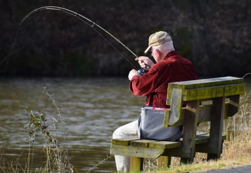 An elderly man in a cap sits on a bench, fishing by a calm lake, focused on his rod.