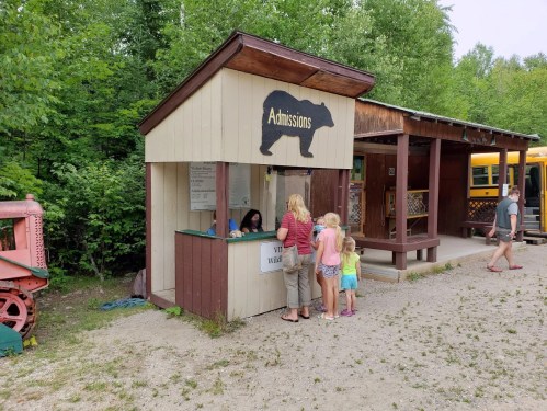 A small admissions booth with a bear silhouette, surrounded by trees, where families are purchasing tickets.