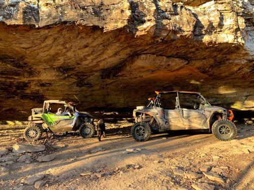 Two off-road vehicles parked under a large rock overhang, with a dog nearby on a dirt path.