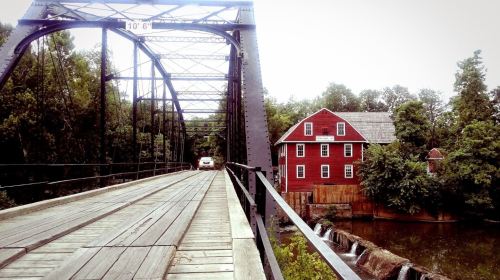 A wooden bridge leads to a red mill house by a river, surrounded by lush green trees.