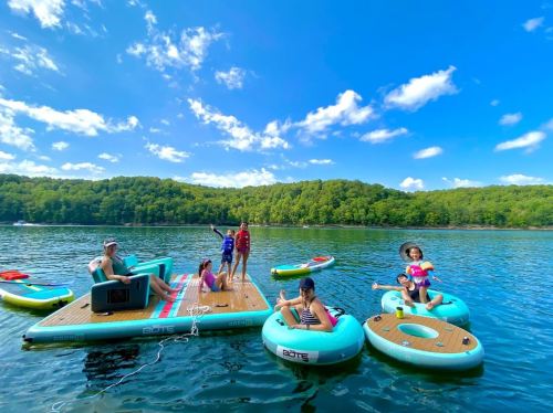 A group of children and adults enjoying a sunny day on floating platforms at a lake, surrounded by greenery.