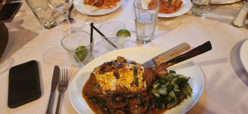 A plate of meat with a bone, topped with cheese and mushrooms, served with greens and drinks on a table.