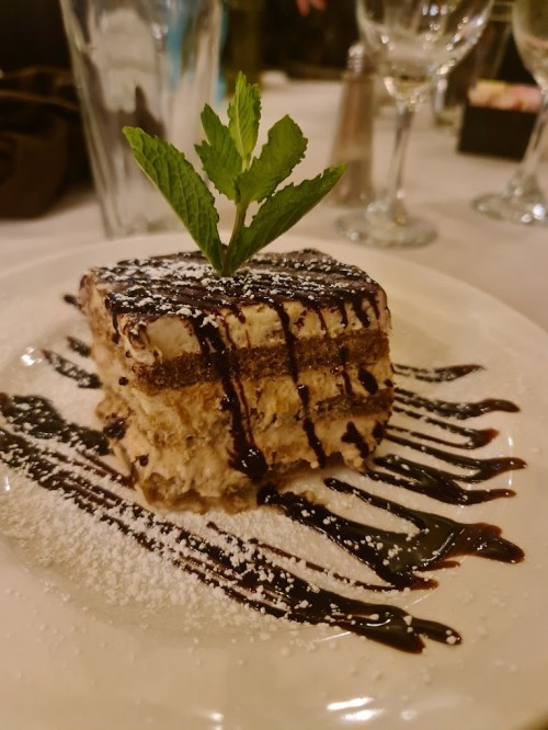 A slice of dessert with layers, drizzled with chocolate sauce and garnished with a mint leaf on a white plate.