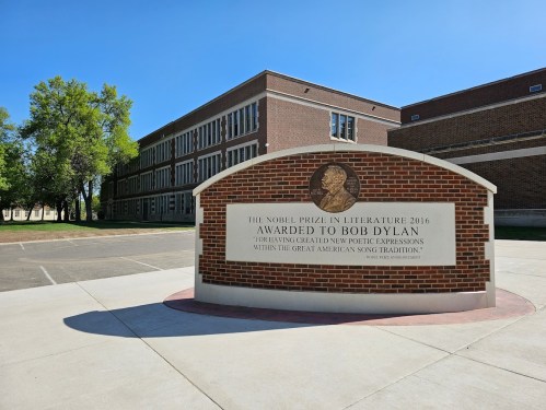 Sign commemorating Bob Dylan's 2016 Nobel Prize in Literature, set against a school building backdrop.