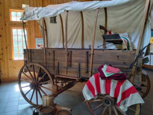 A vintage wooden wagon with a white canopy, displayed indoors, featuring a colorful blanket and wooden wheels.