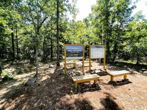 Information sign at Blue Mountain trailhead, surrounded by trees, with benches for visitors to sit.