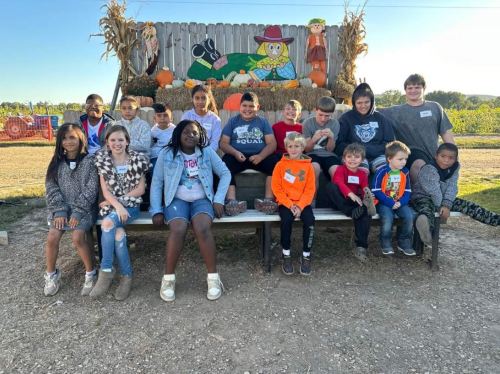 A group of children and teens sitting on a bench in front of a festive fall backdrop with pumpkins and decorations.