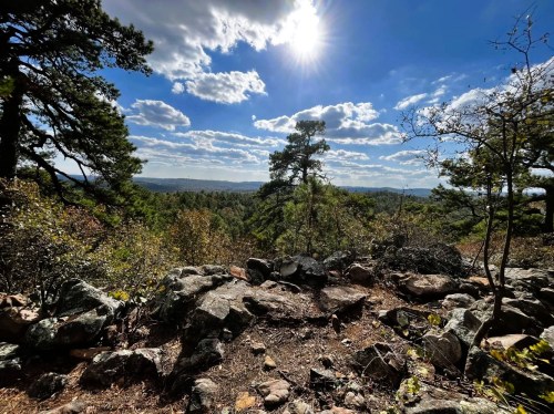 A scenic view of a rocky landscape with trees under a bright blue sky and fluffy clouds, sun shining in the background.