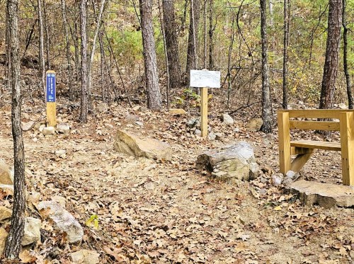 A wooded area with a trail marker, a sign, and a wooden bench surrounded by rocks and fallen leaves.