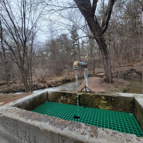 A water fountain with a spout, surrounded by trees and a creek, on a cloudy day.