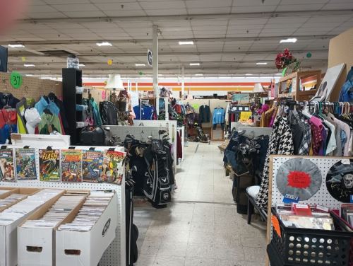 A wide view of a thrift store aisle filled with clothing racks, comic books, and various items for sale.