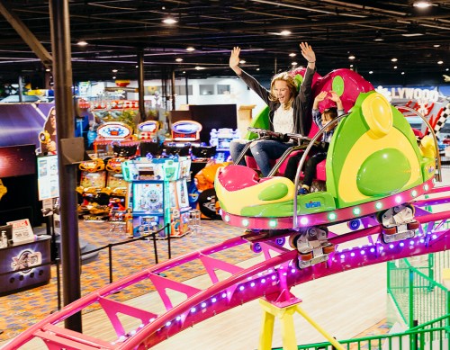 A woman and child joyfully ride a colorful indoor roller coaster in an arcade filled with games.