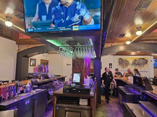 A restaurant interior featuring a bar, hanging glasses, and a TV displaying a sports game, with patrons seated at tables.