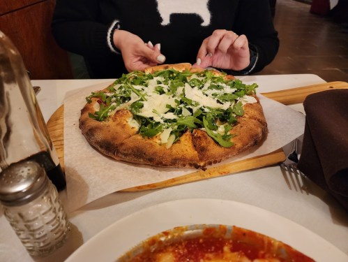 A person is preparing to eat a pizza topped with arugula and cheese, with a side dish of pasta in the foreground.