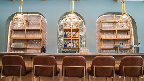 A stylish bar with a light blue wall, gold accents, and shelves displaying various bottles and glassware.